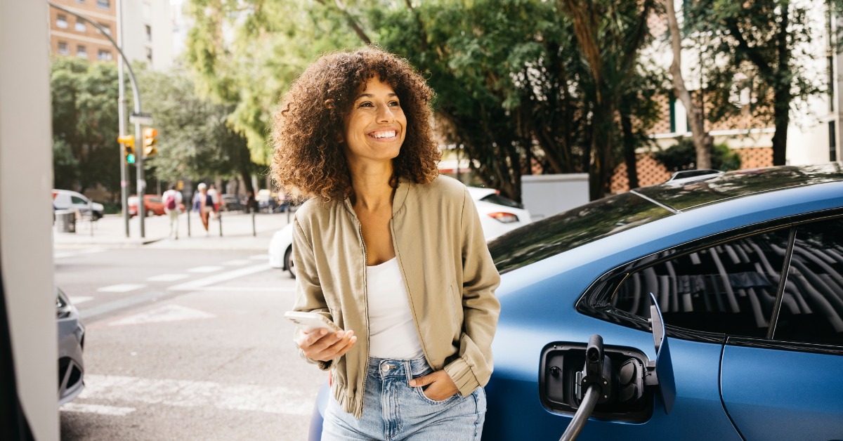 A person holding a cellphone smiles while leaning against their blue electric vehicle attached to a charger.