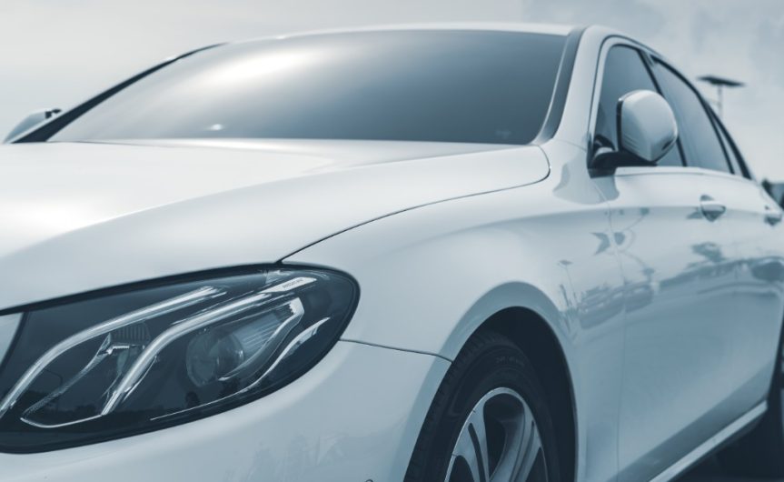 A white sedan parked outdoors with a prominent front headlight, dark windows, side mirror, and front wheel visible.