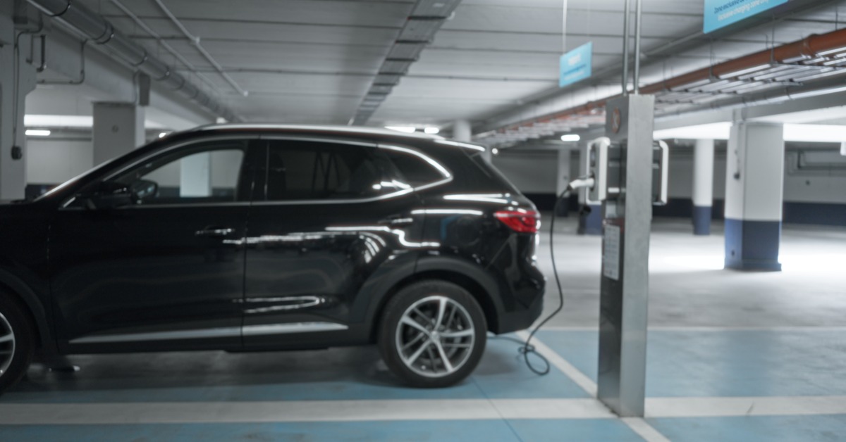 A black SUV parked beside an electric charging station in an underground garage with concrete pillars and ceiling lights.