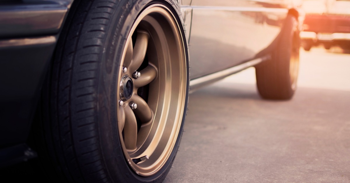 A black tire with a bronze five-spoke alloy wheel on a silver vintage car parked on pavement in warm sunlight.