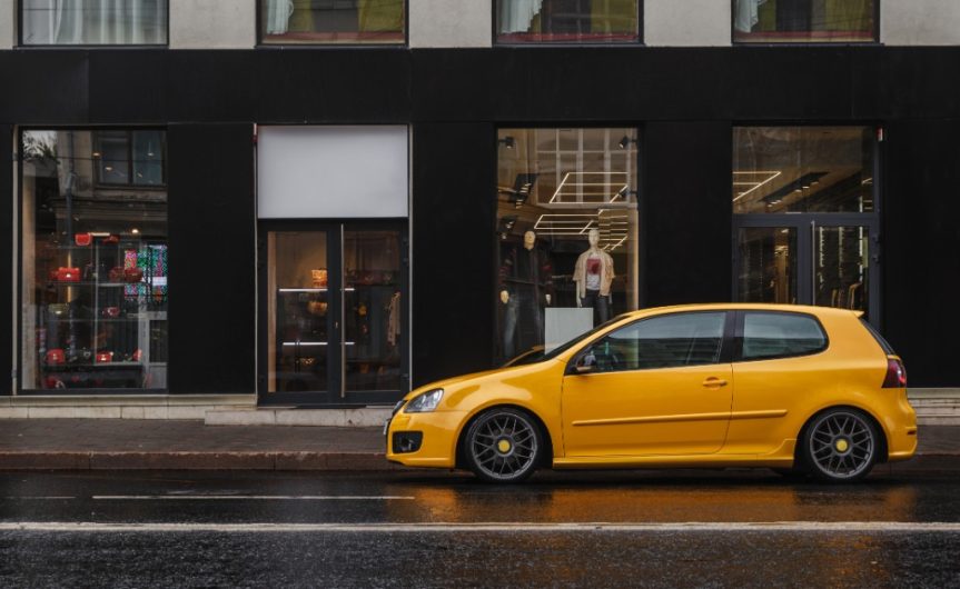A small yellow car parked on a city street during the daytime. The car is next to a large clothing store.