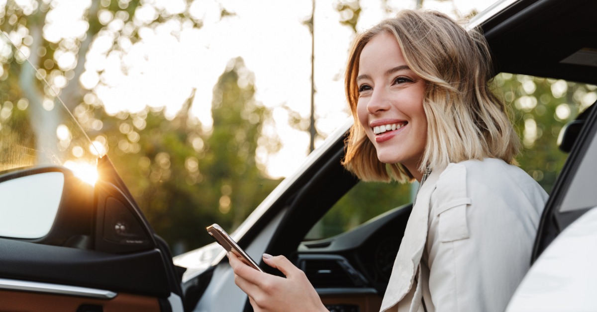 A person smiling and looking at their phone while sitting in a parked car with the door open during the daytime.