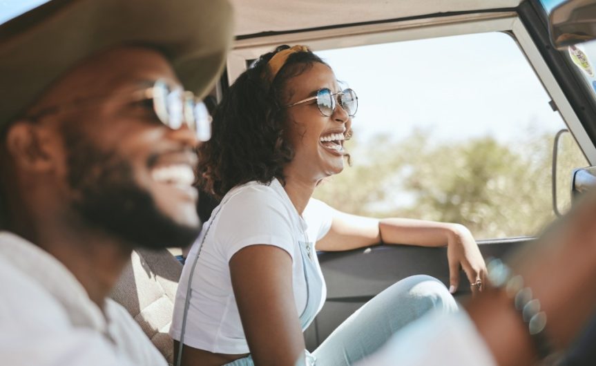 Two people wearing sunglasses in the front seats of a vehicle on a sunny day. Both people are laughing in the car.