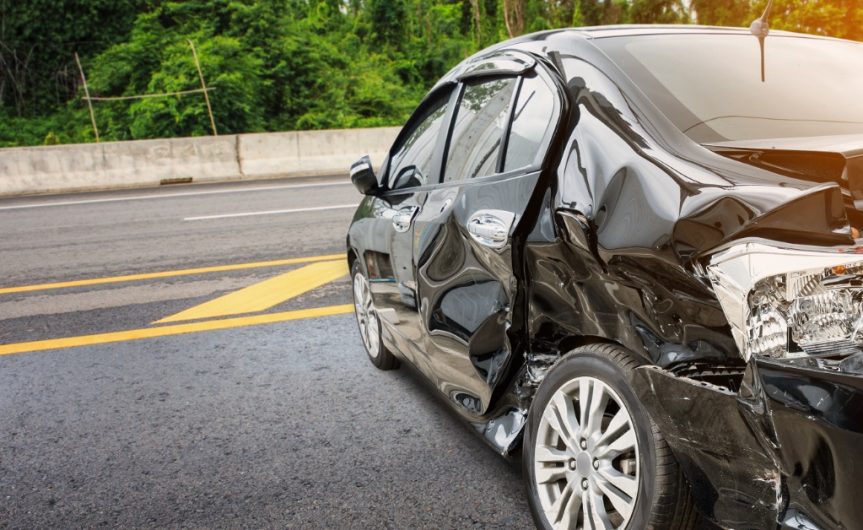A black car sideways on the side of the road with significant crash damage to its side exterior and taillights.