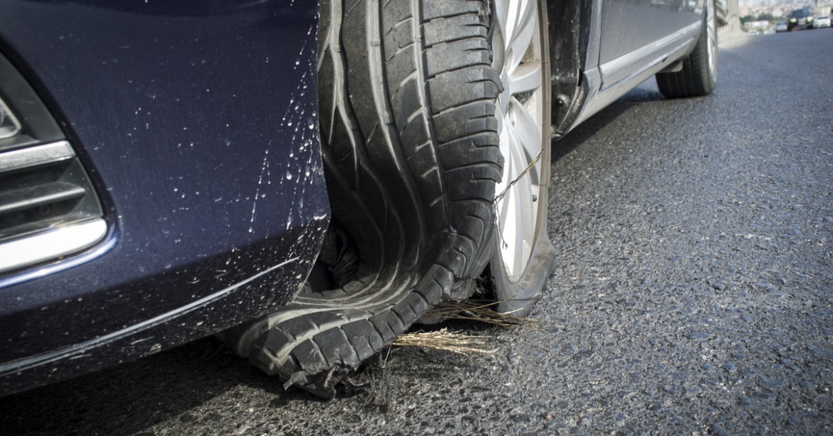 A close-up of the ruptured tire of a ble car on the a pved road with the tread disconnected and plies exposed.