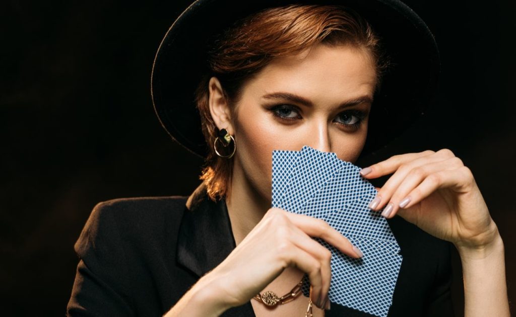 Attractive woman in jacket and hat covering face with poker cards isolated on black background