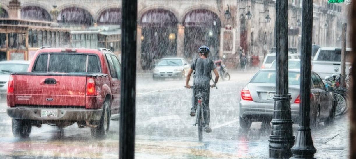 Carro rojo y persona en bicicleta en medio de fuertes lluvias. Ilustra nota sobre consejos para prevenir inundaciones por lluvias.