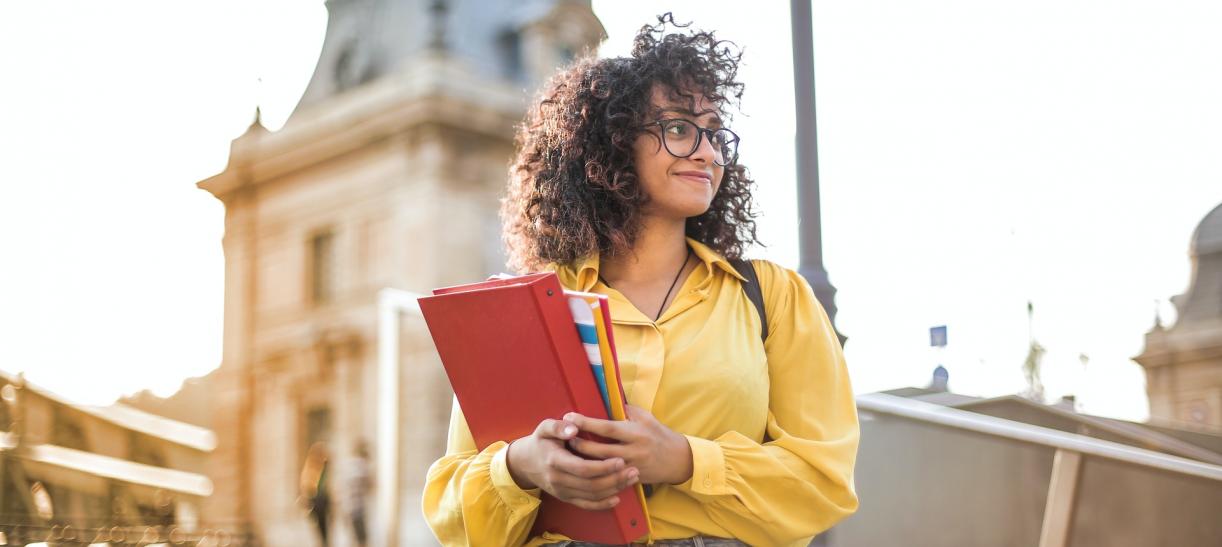 Foto de joven mujer llevando libros y carpetas consigo, con una iglesia a sus espaldas.