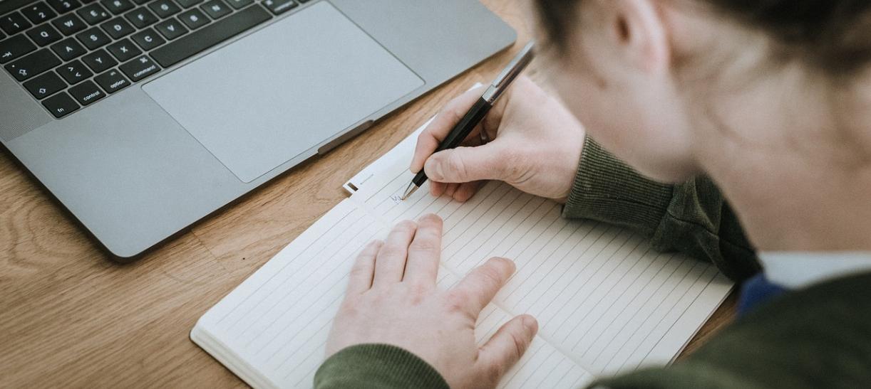 Mujer joven escribiendo en un cuaderno con un computador en frente.