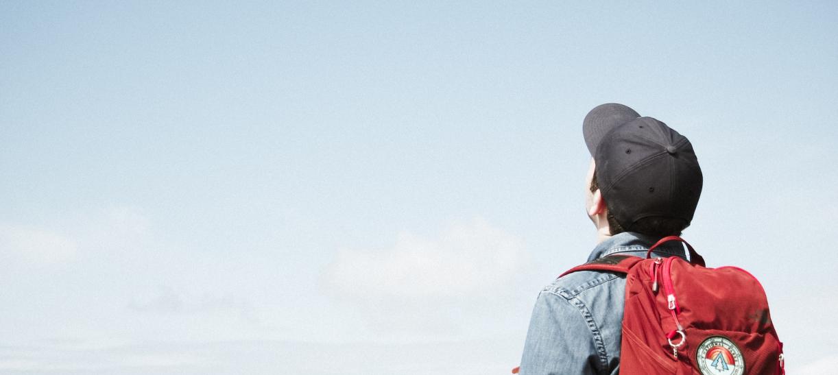 Jóven con mochila y gorra mira hacia el cielo. Ilustra nota sobre becas.