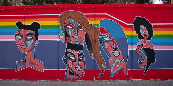 Mural painted on a wall. It's a colourful illustration of 5 faces, all different. Behind them there are rainbow stripes from one side to the other.
