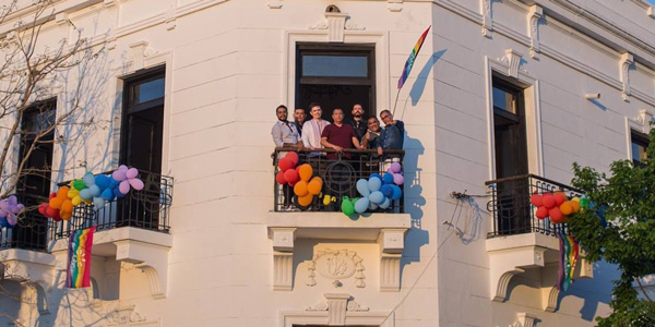Seven people stand on a second floor balcony. One of them holds a rainbow flag. The other balconies are decorated with balloons in rainbow colours.