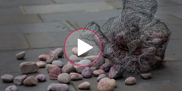 The image shows the sculpture surrounded by rocks on the sidewalk.