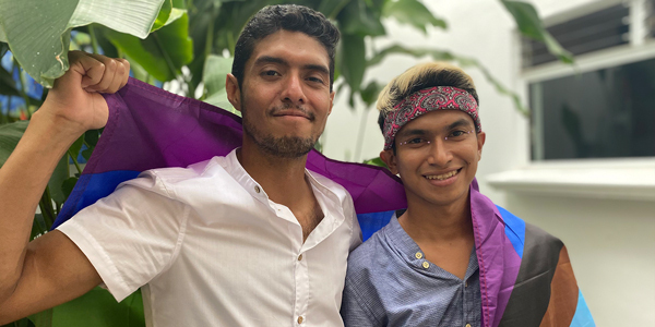 The image shows two people with male gender presentation. The person on the left has dark hair and brown skin wearing a white shirt and smiling slightly. On the right is the other person who is smiling, showing her teeth, wears a scarf on her forehead, and has light colored hair. Both people are holding a rainbow flag.
