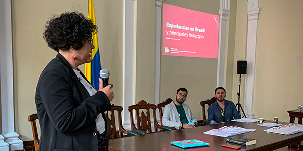 Ana from All Out presents information in front of a screen projection that says Experiences in Brazil and Main Findings in Spanish.