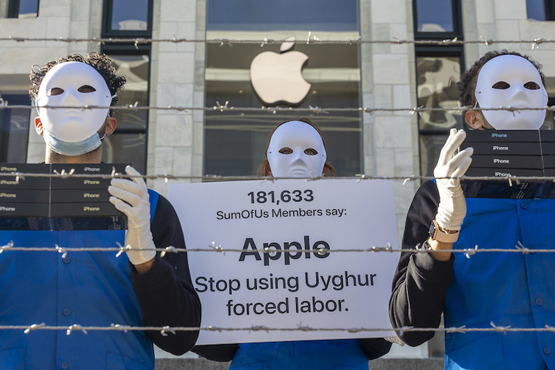 Three people facing the camera in plain white carnival mask with a neutral expression covering the whole face with cut outs for eyes They are wearing blue tops with black sleeves The two people either side are holding boxes of apple products with gloved hands There is a banner between them reading 181633 SumOfUs member said Apple stop using Uyghur forced labour They are standing behind five strands of barbed wire in front of an Apple store with the company logo prominently displayed