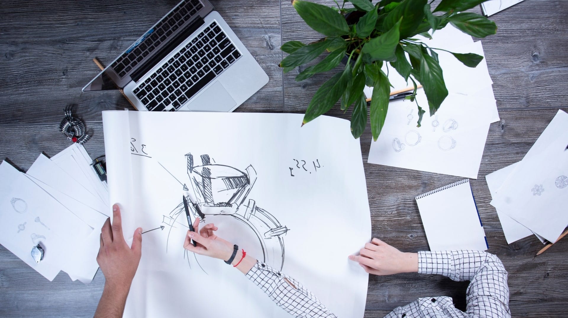 Jewelry designer working on a detailed ring sketch on a large sheet of paper surrounded by tools, plants, and a laptop.