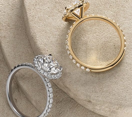 Two engagement rings in gold and platinum, featuring pave-set diamonds and round-cut centers, displayed on a neutral stone backdrop.