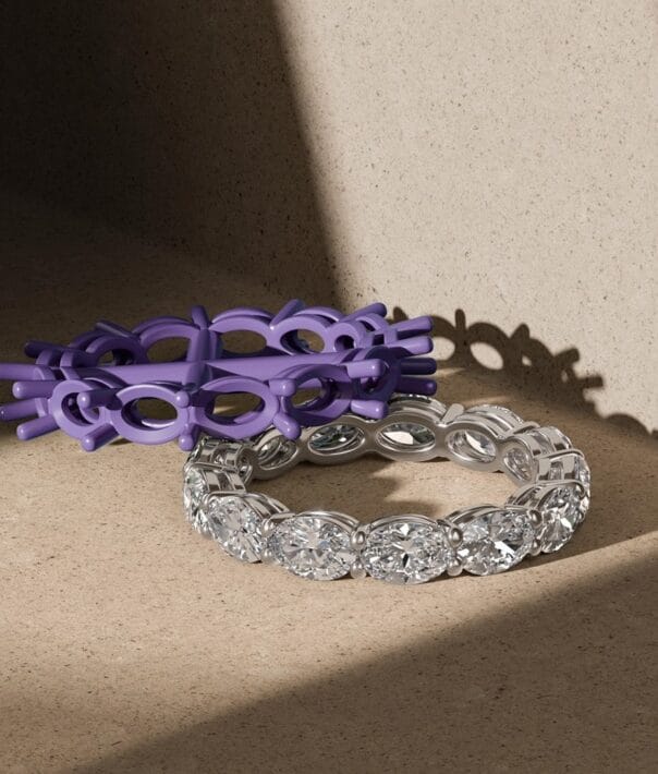 Oval-cut diamond eternity ring next to a purple plastic ring model on a textured beige surface under natural lighting