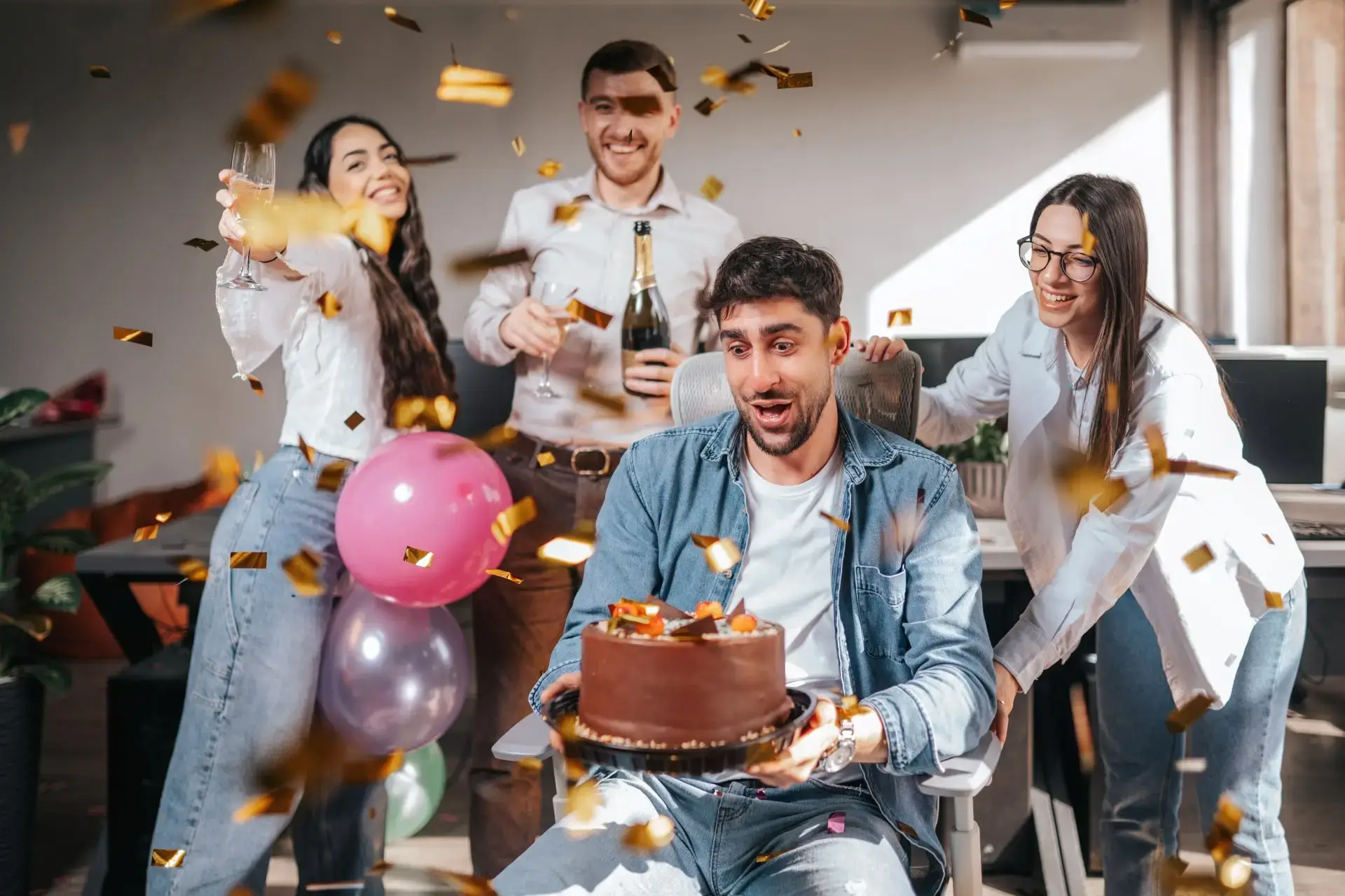 A group of office workers celebrating a birthday with a cake, balloons, and confetti.