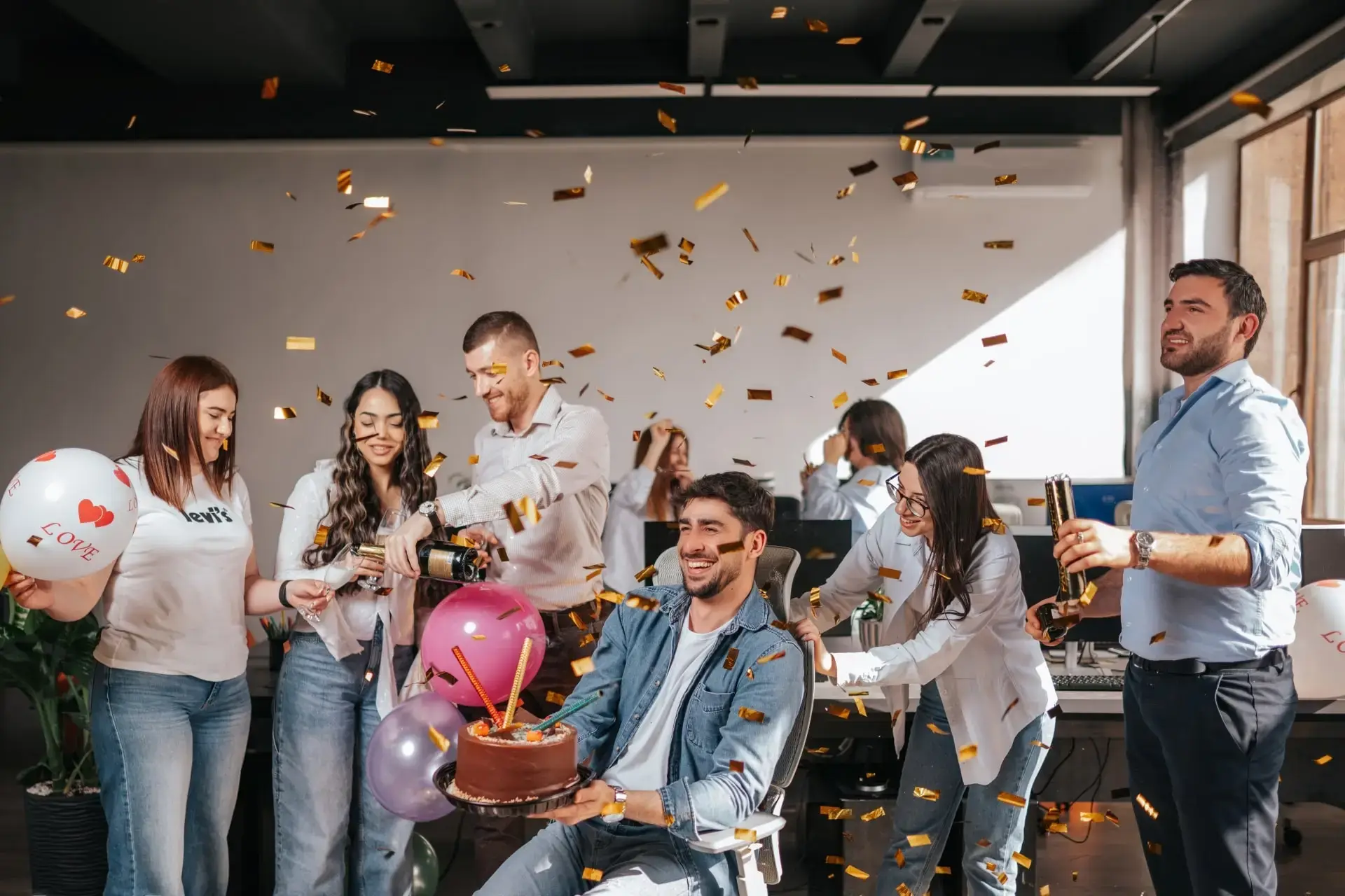 A group of people celebrating a birthday in an office with balloons, cake, and confetti.