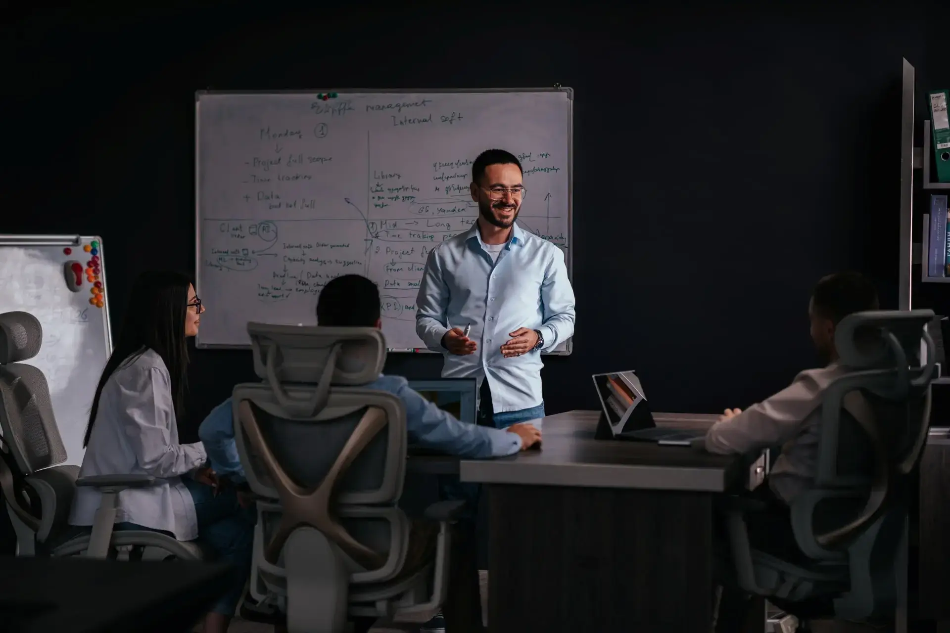 A man is presenting to a group of people seated around a table in an office setting with a whiteboard in the background.