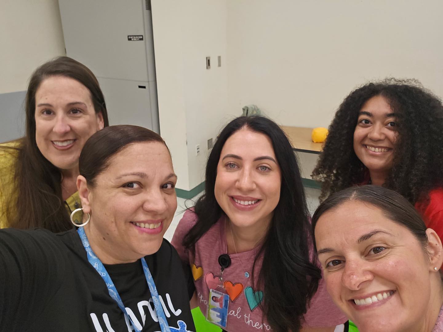 Five smiling women pose for a selfie indoors.