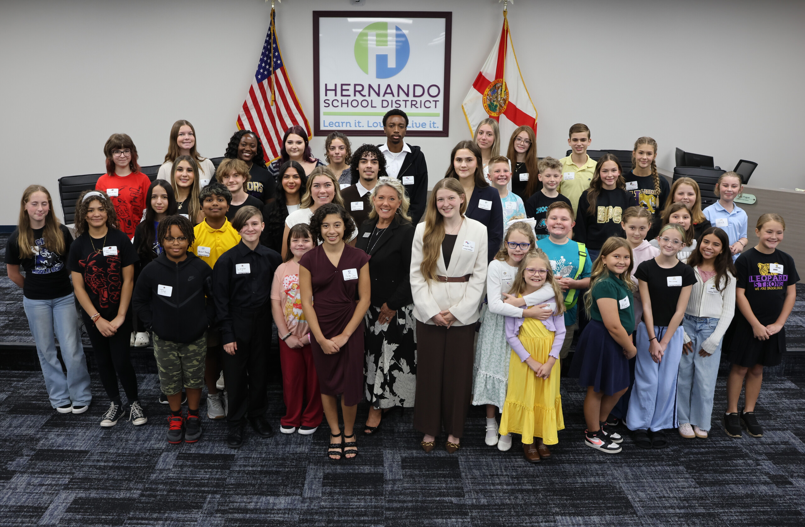 A large group of students and adults pose together indoors for a photo in front of flags and a sign.