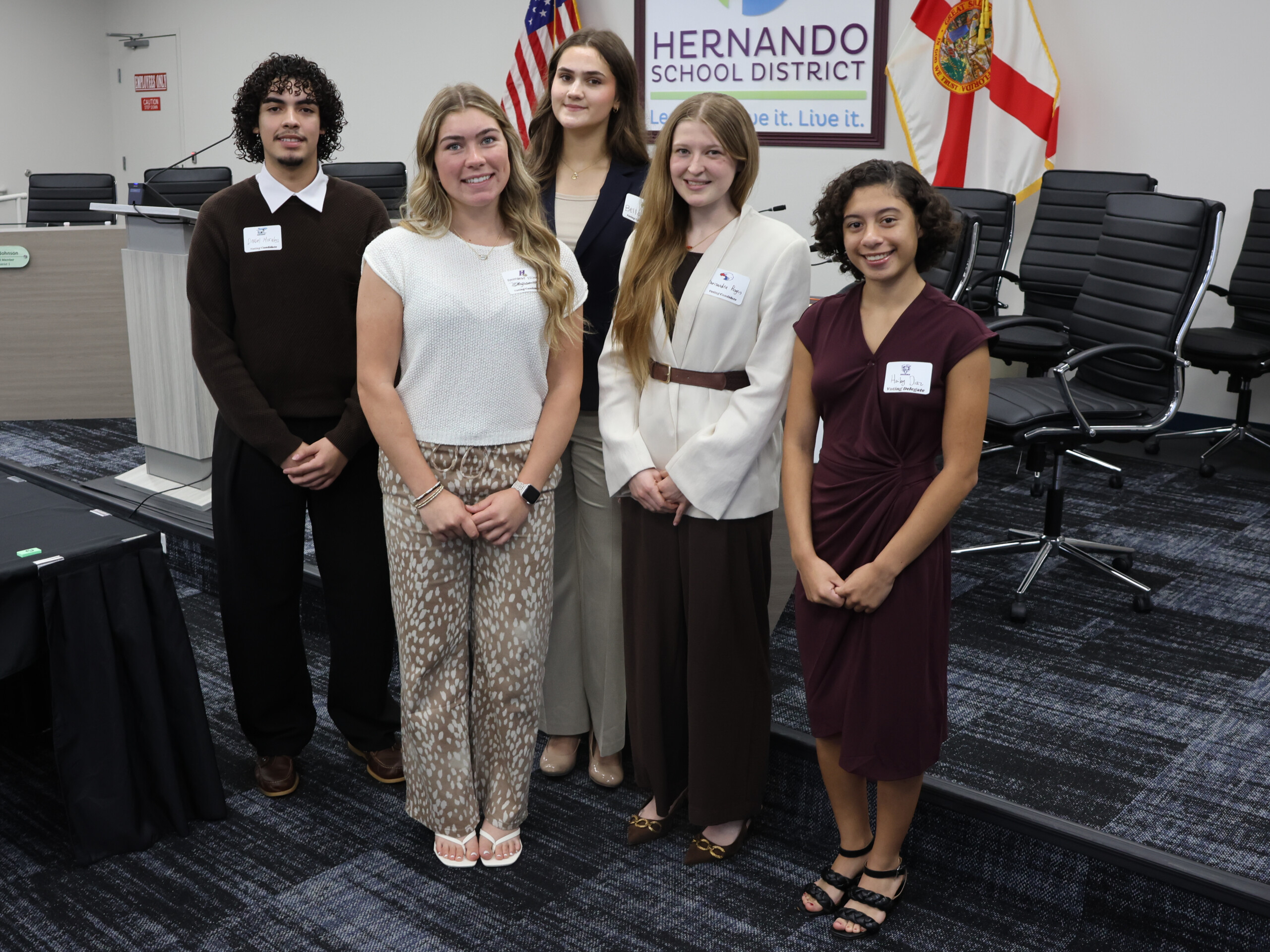 Five young people stand together smiling in a meeting room with flags and a sign reading 'Hernando School District'.