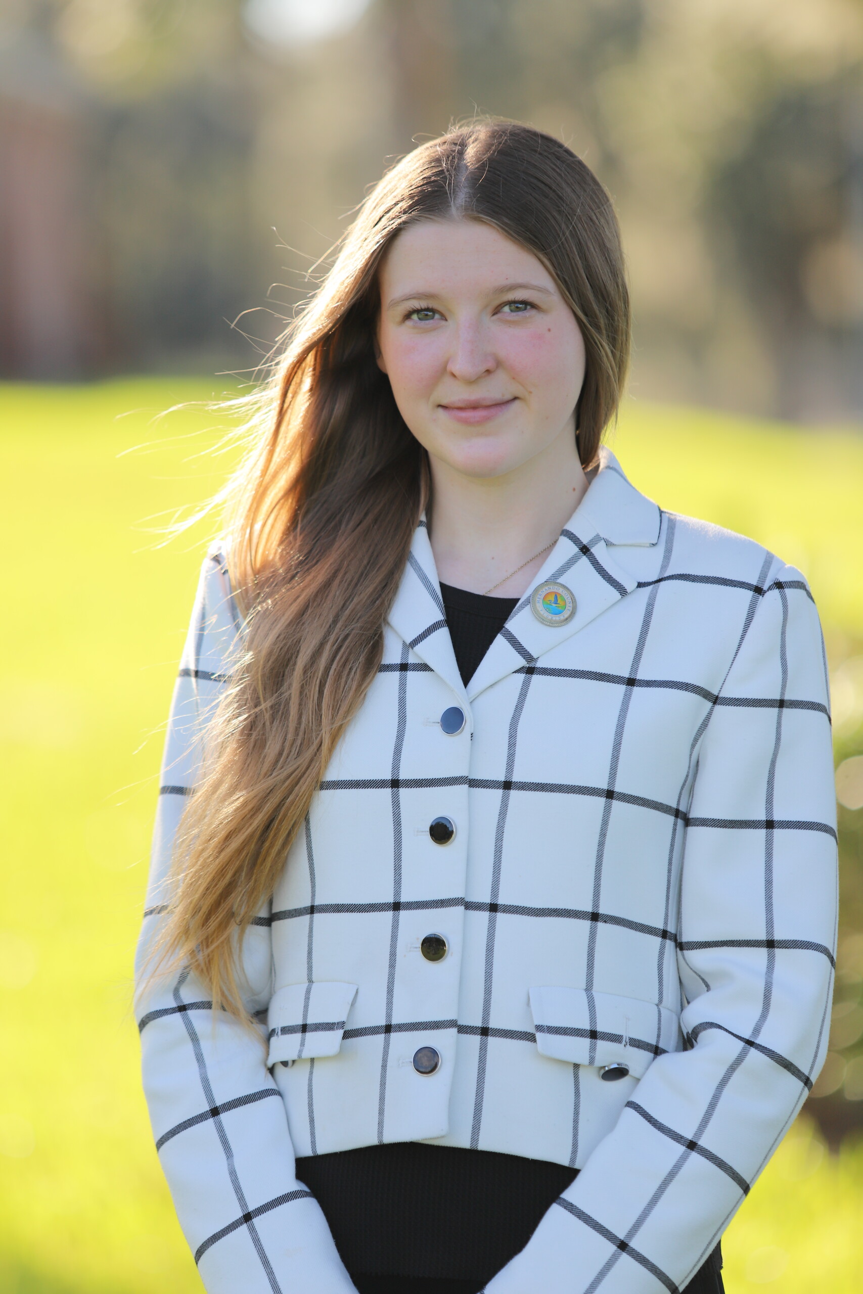 A young person with long brown hair smiles outdoors while wearing a white plaid blazer.
