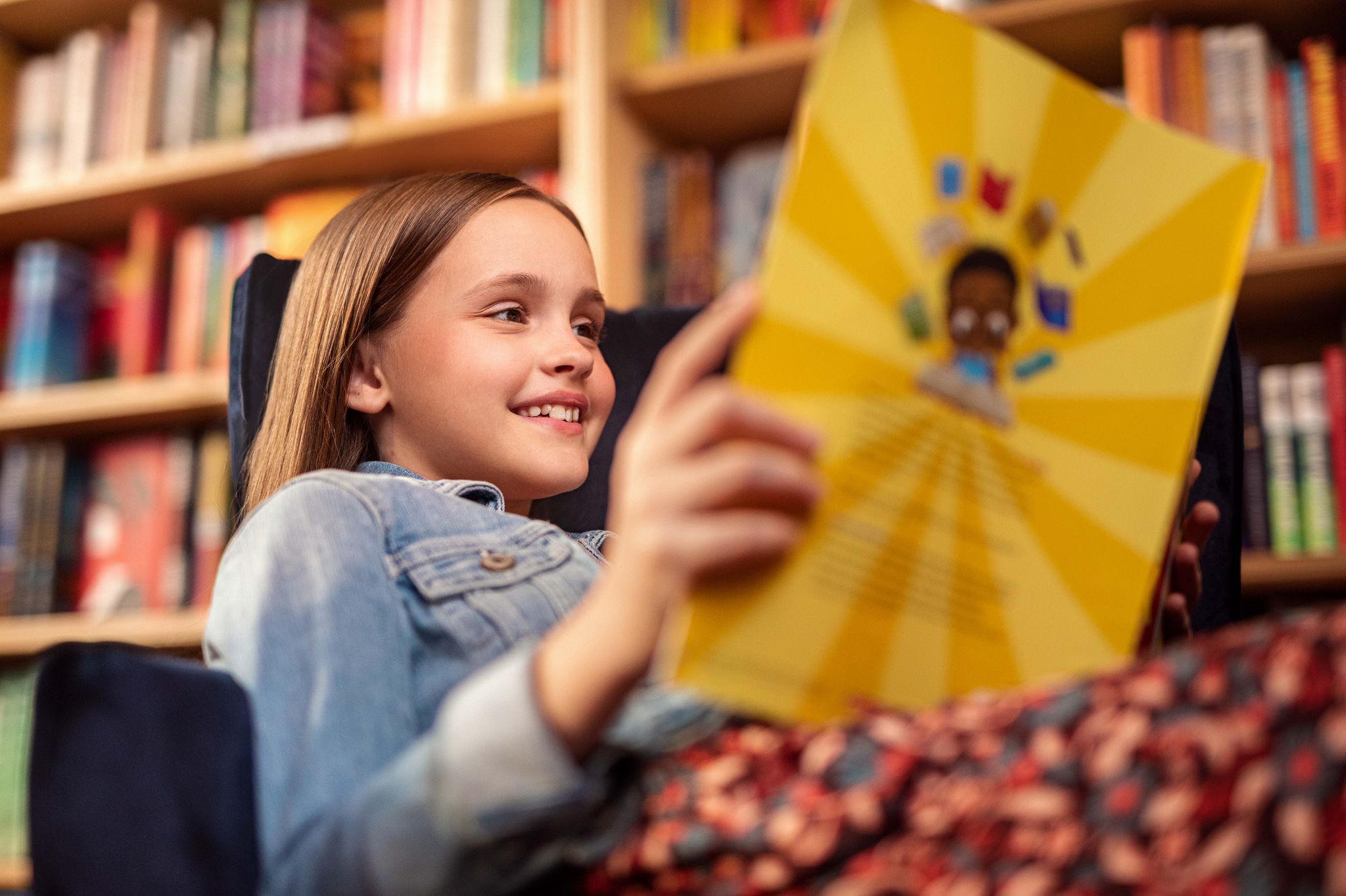 A young girl smiles while reading a colorful book in a library, with bookshelves in the background.