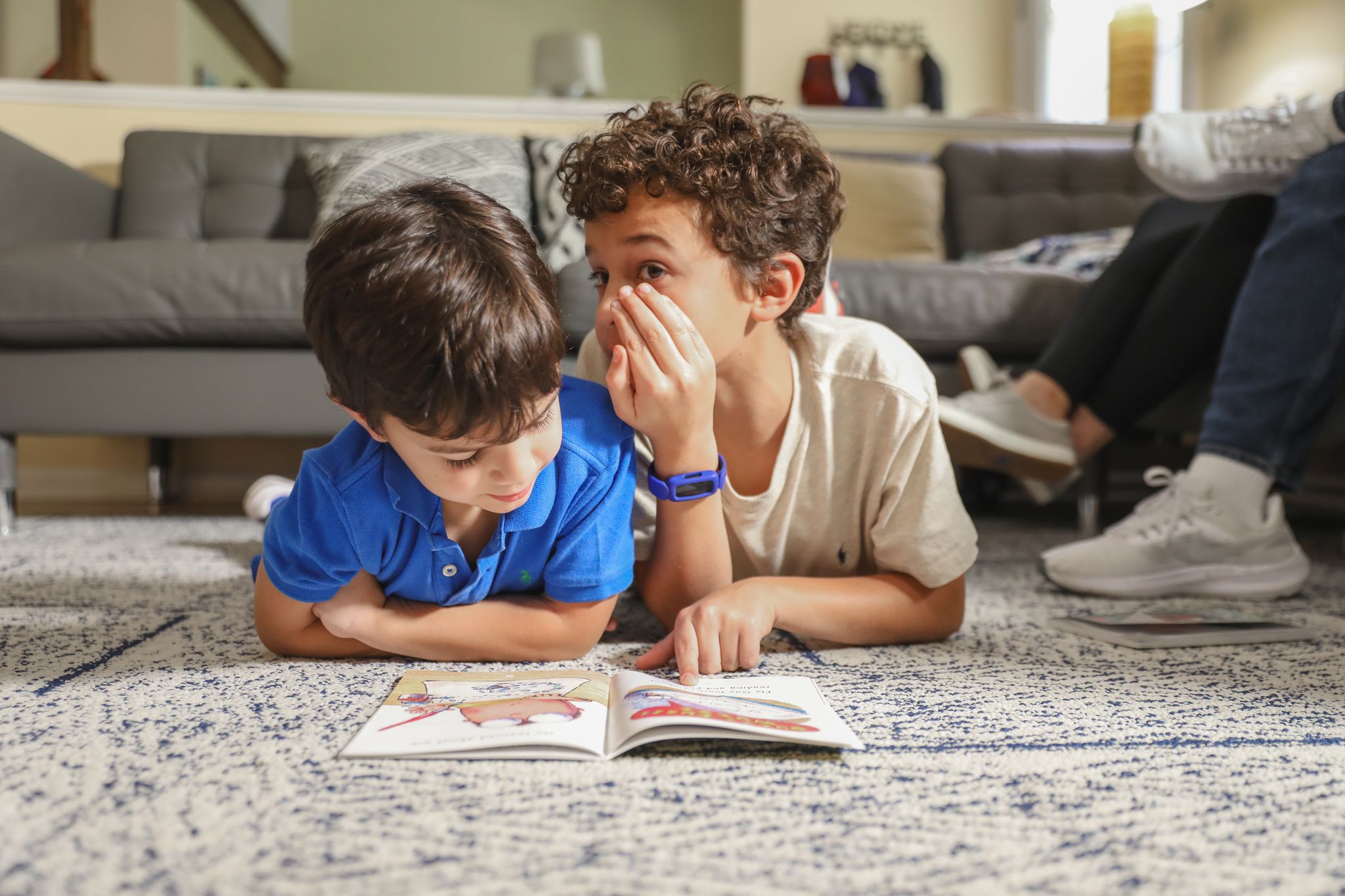 Two young boys whisper secrets to each other while looking at a book on the floor.