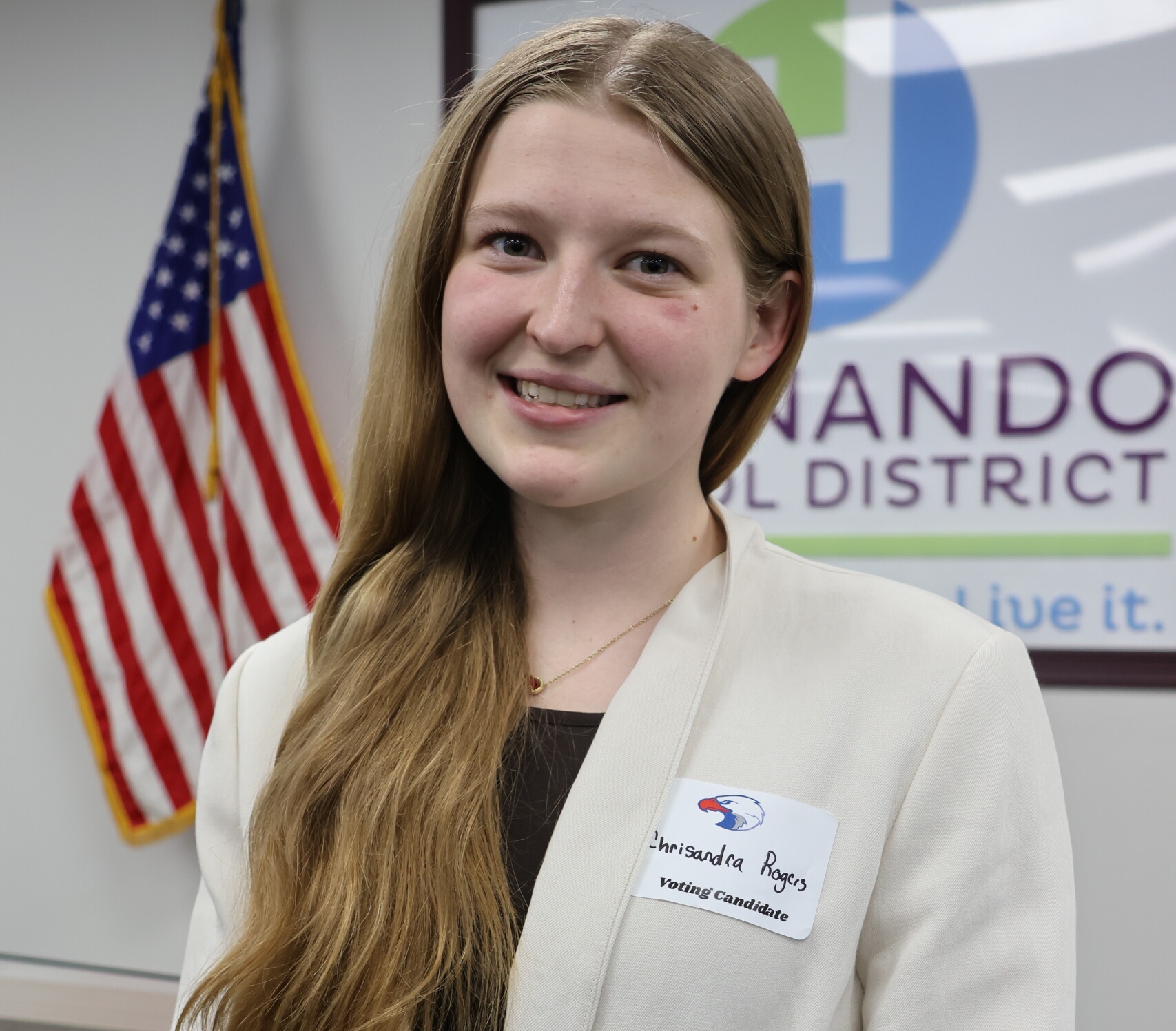 A young woman with long blonde hair smiles in an office, wearing a white blazer and a name tag that reads "Chrisandra Rogers, Voting Candidate".