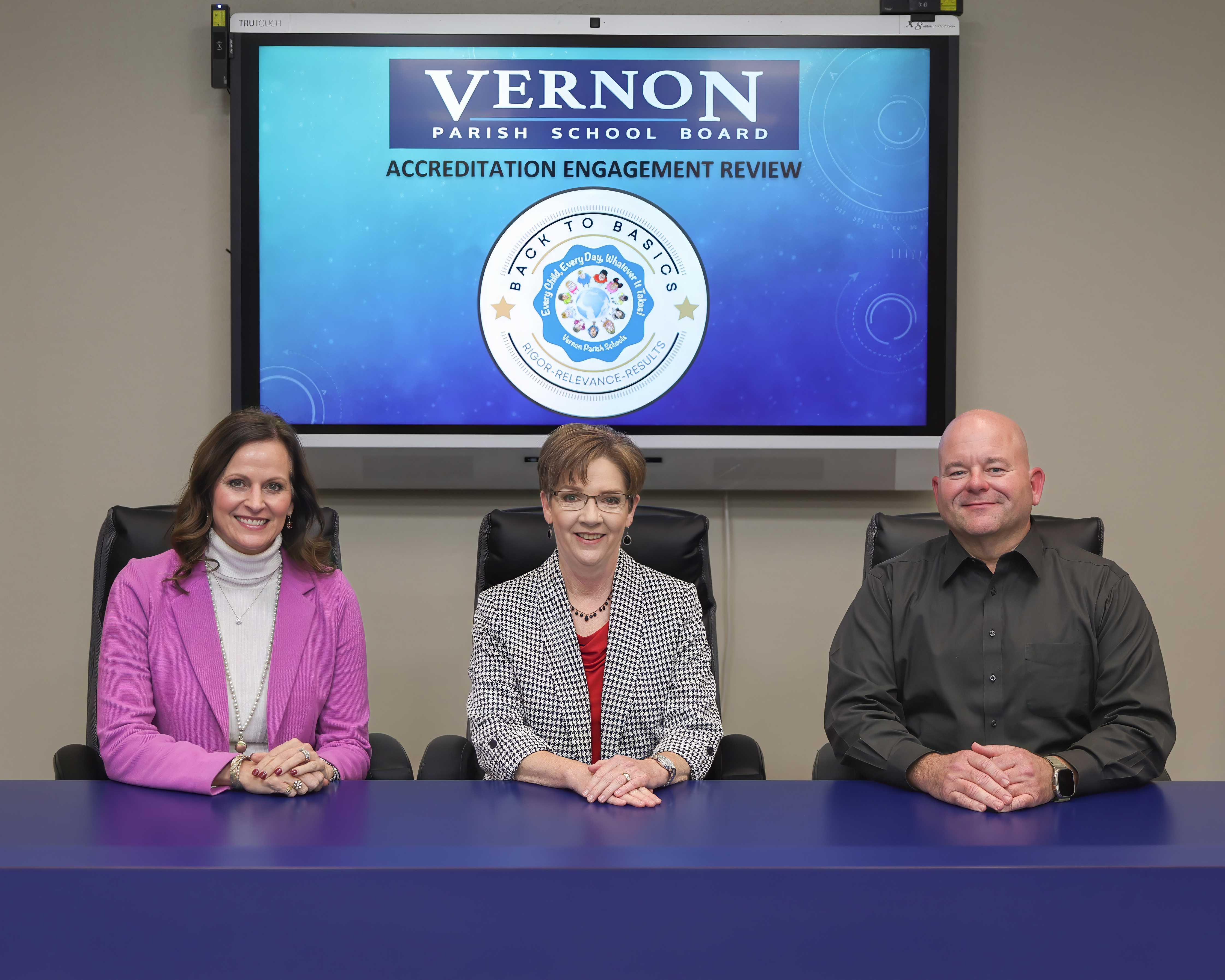 Three people sit at a table in front of a screen with the word 'Vernon' displayed.