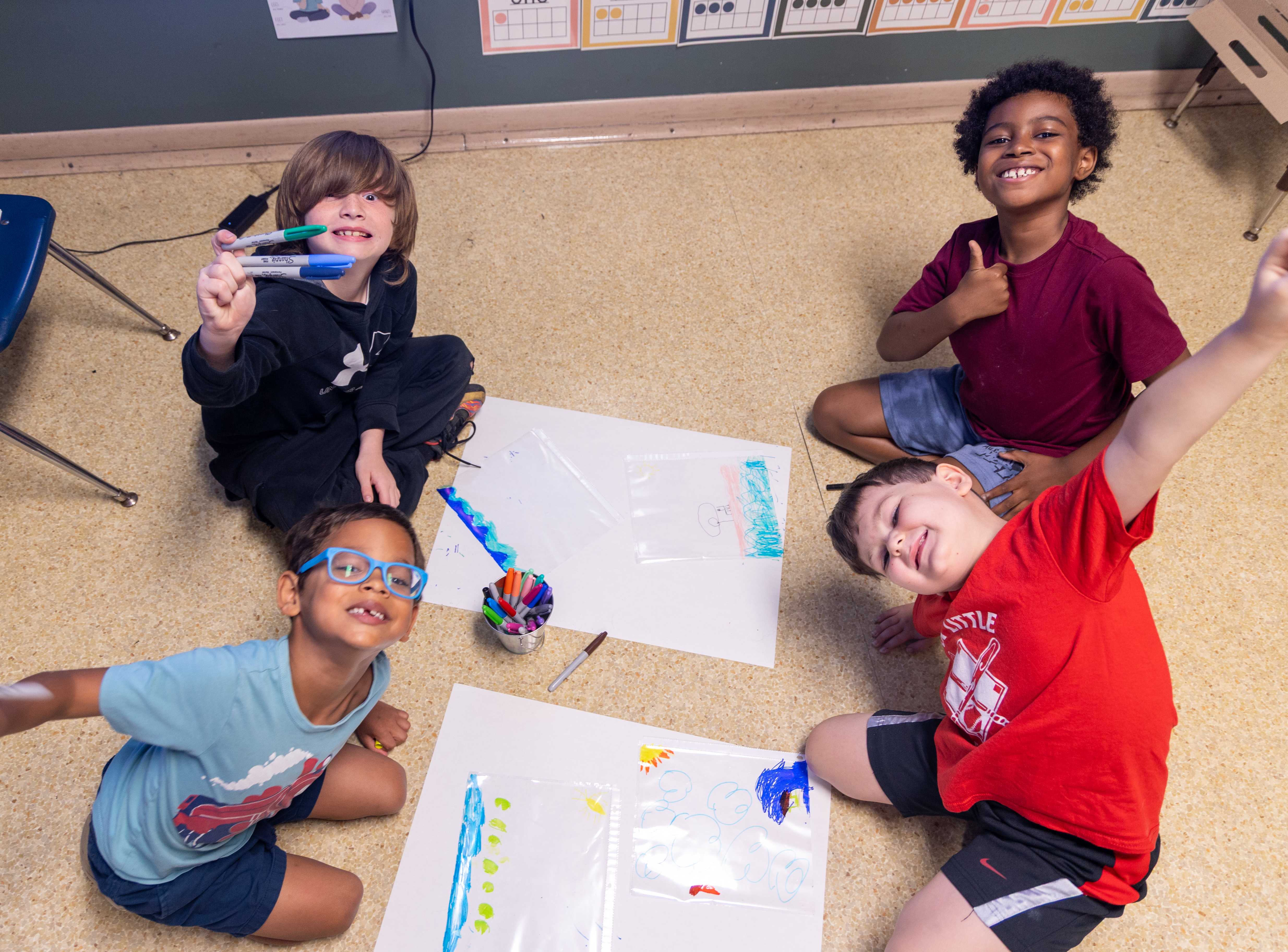 Four smiling children sit on the floor around art projects, holding up their creations.