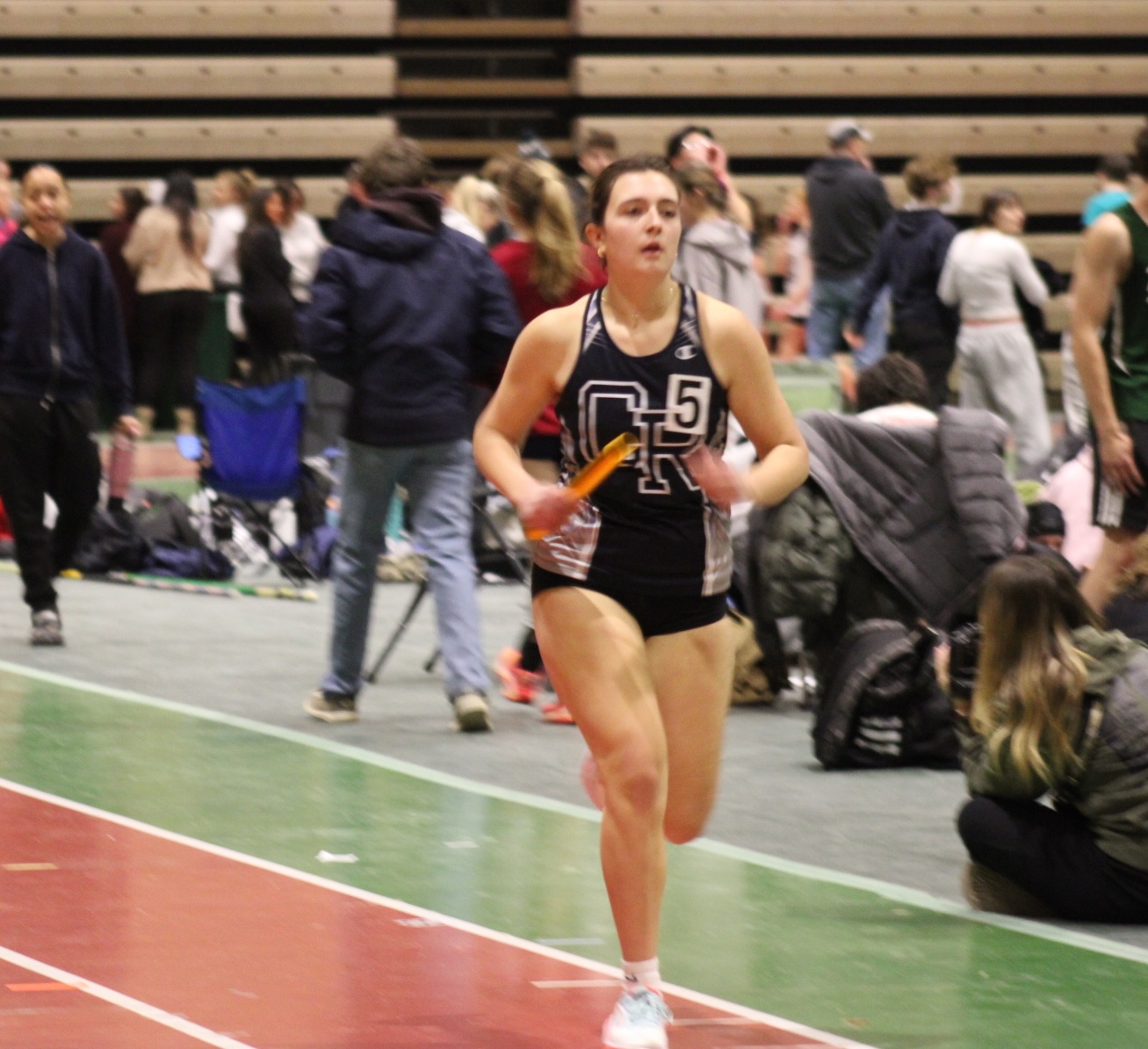 A female athlete runs on an indoor track, holding a baton during a relay race.