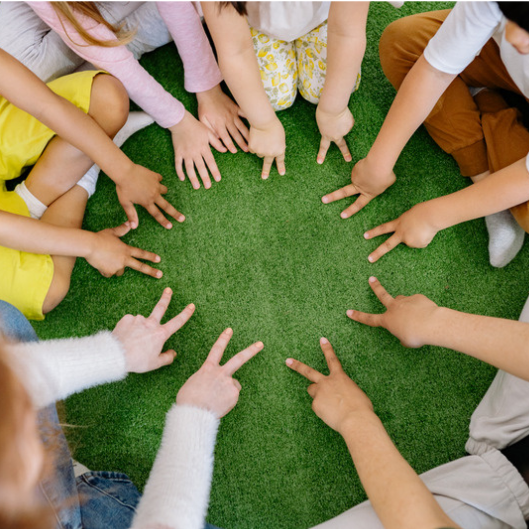 A group of children sit in a circle on green turf, pointing inward toward the center.