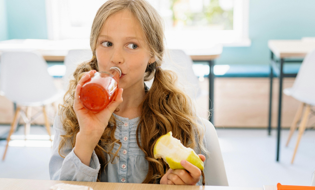 A young girl sips from a juice bottle while holding a half-eaten apple in her other hand.