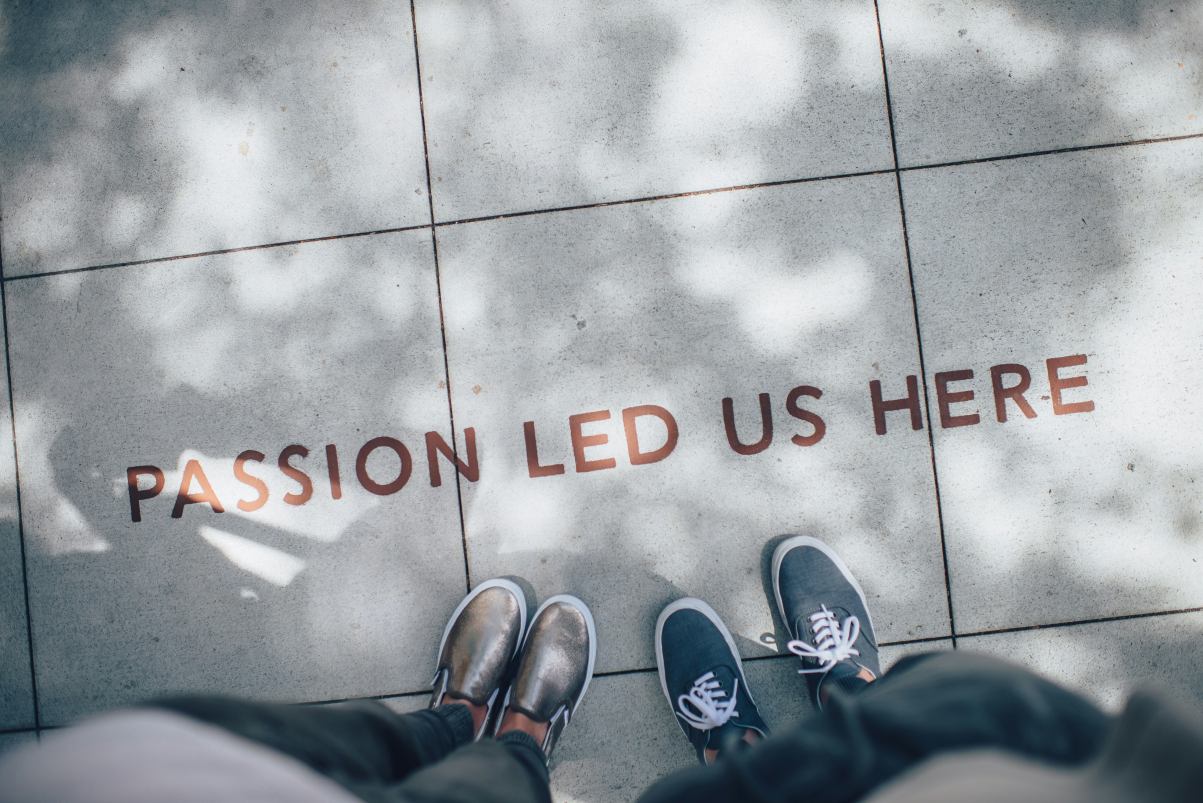 Two pairs of feet stand on a tiled surface with the words 'PASSION LED US HERE'.