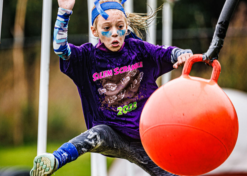 A young girl with face paint leaps over an obstacle during an outdoor race.