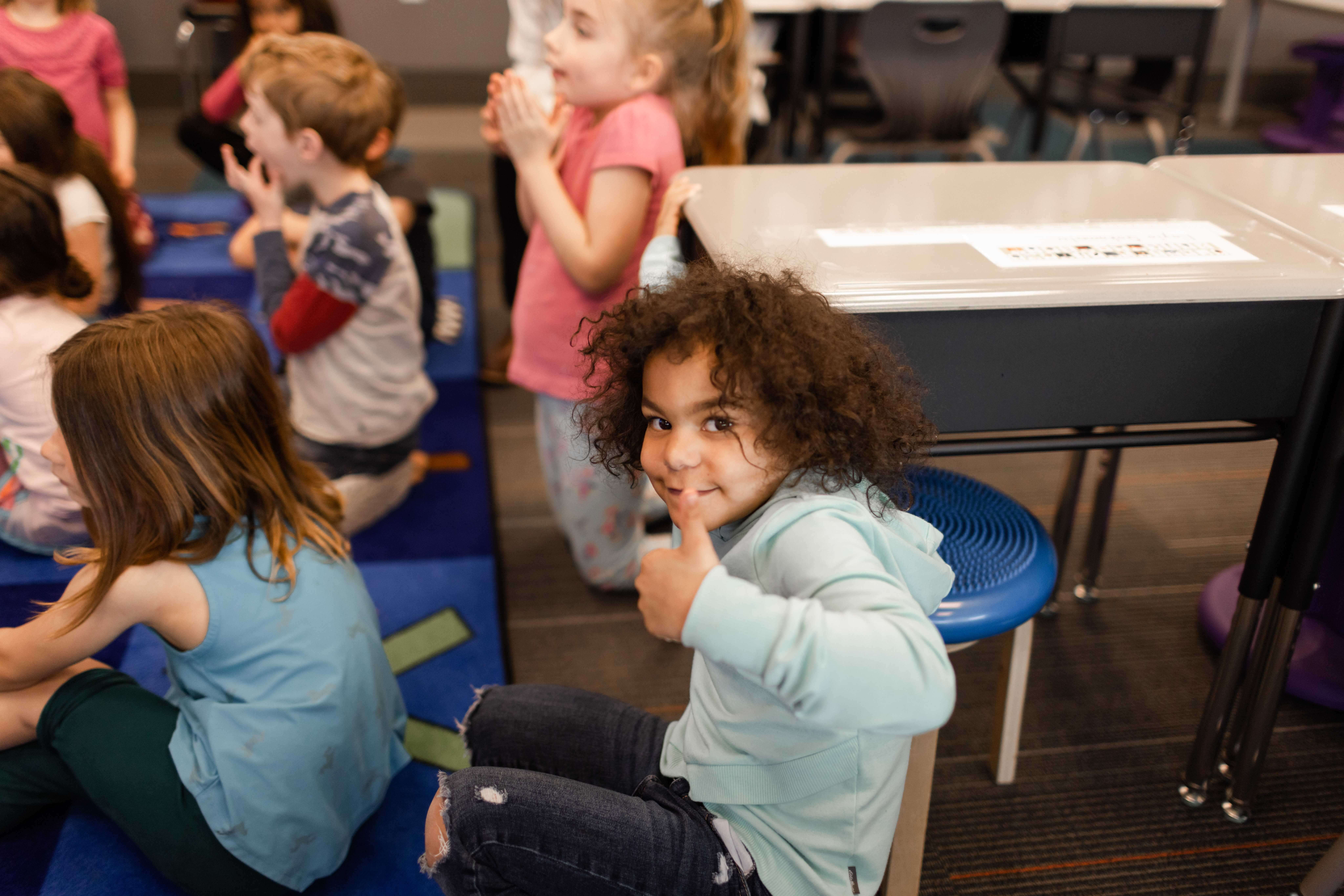 A young girl with curly hair smiles and gives a thumbs up.