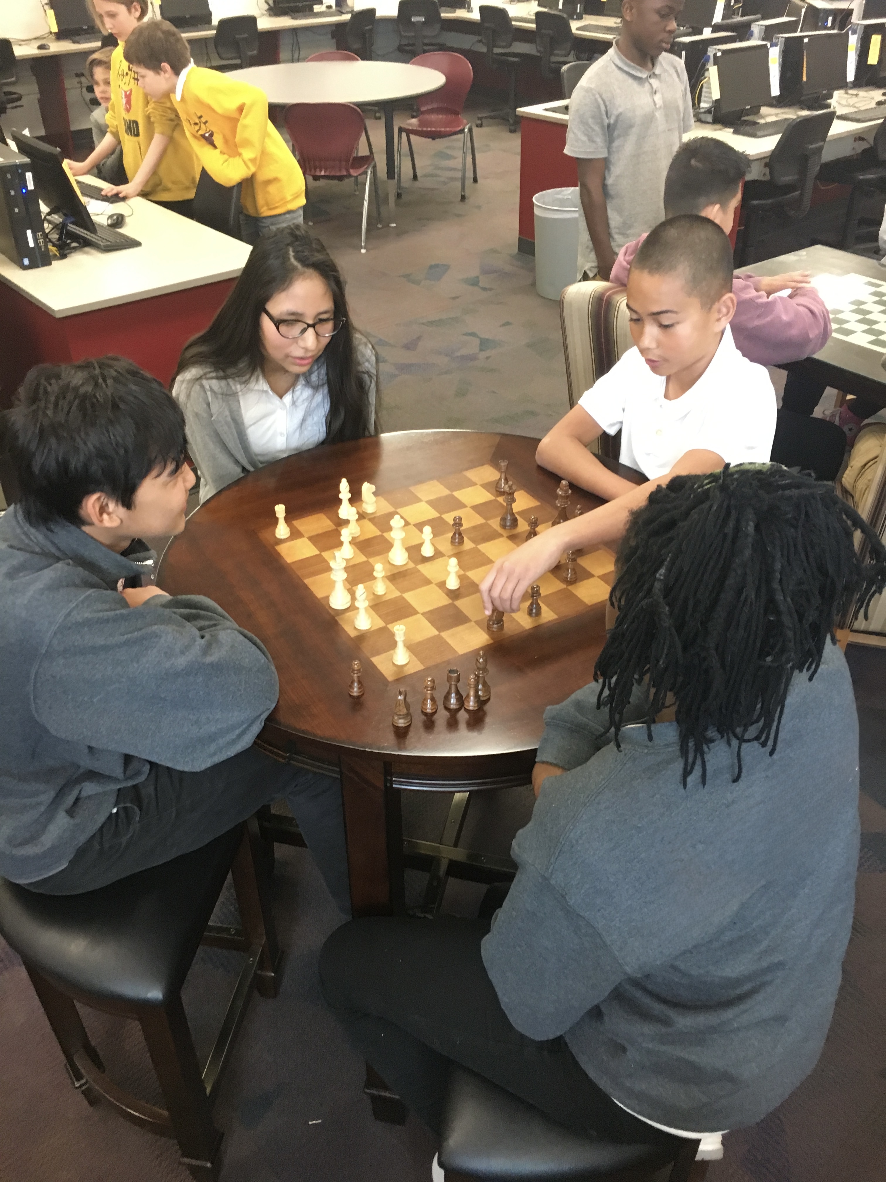 Four students gather around a round wooden table to play a game of chess in a school room.