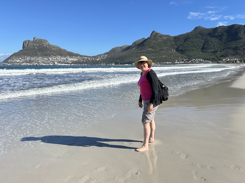 A person stands on a sandy beach, looking towards the camera with mountains in the background.