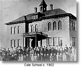 A vintage black and white photo shows a large brick school building with a group of students and teachers.