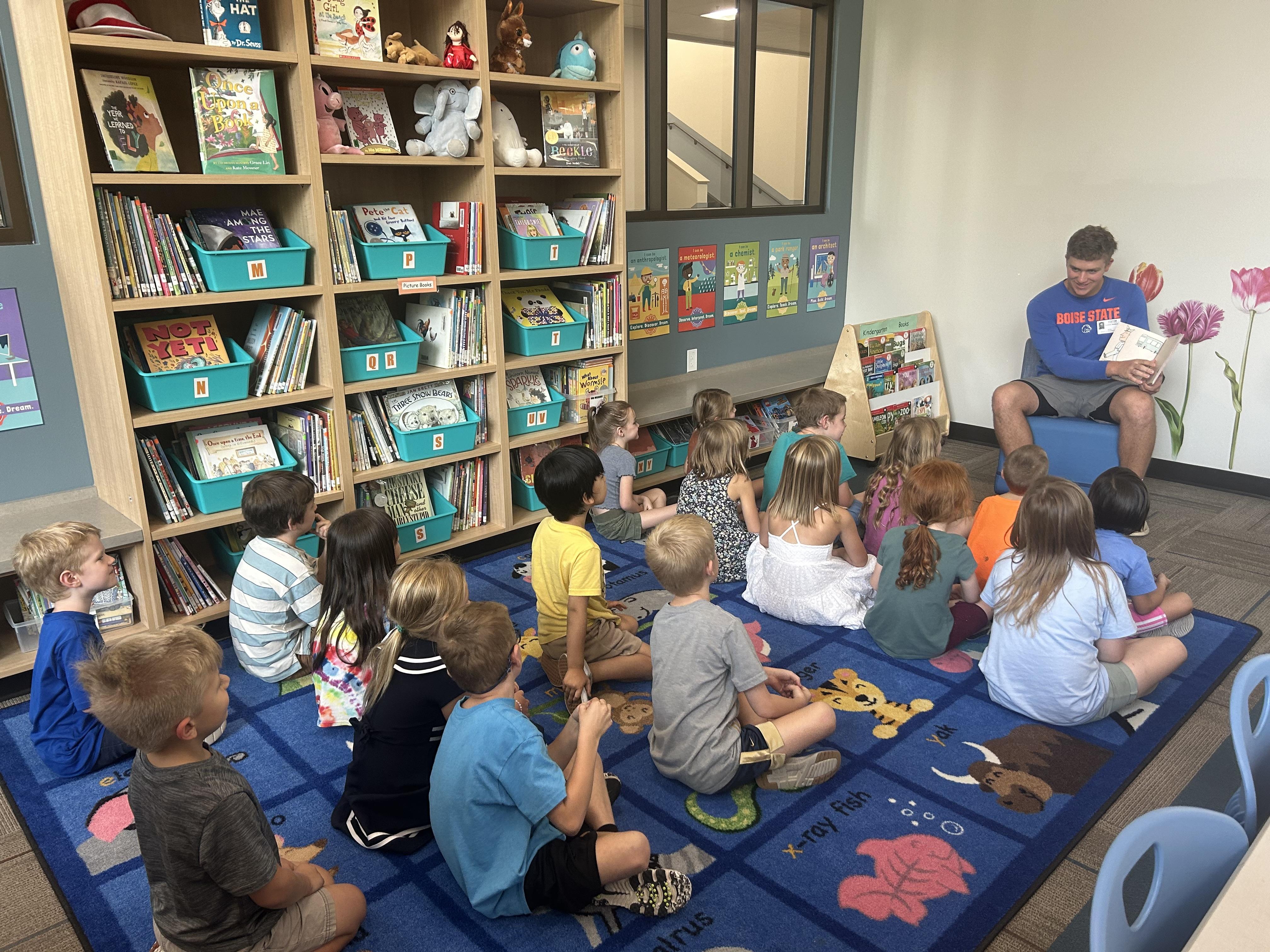 A teacher reads to a group of children seated on mats in a library.