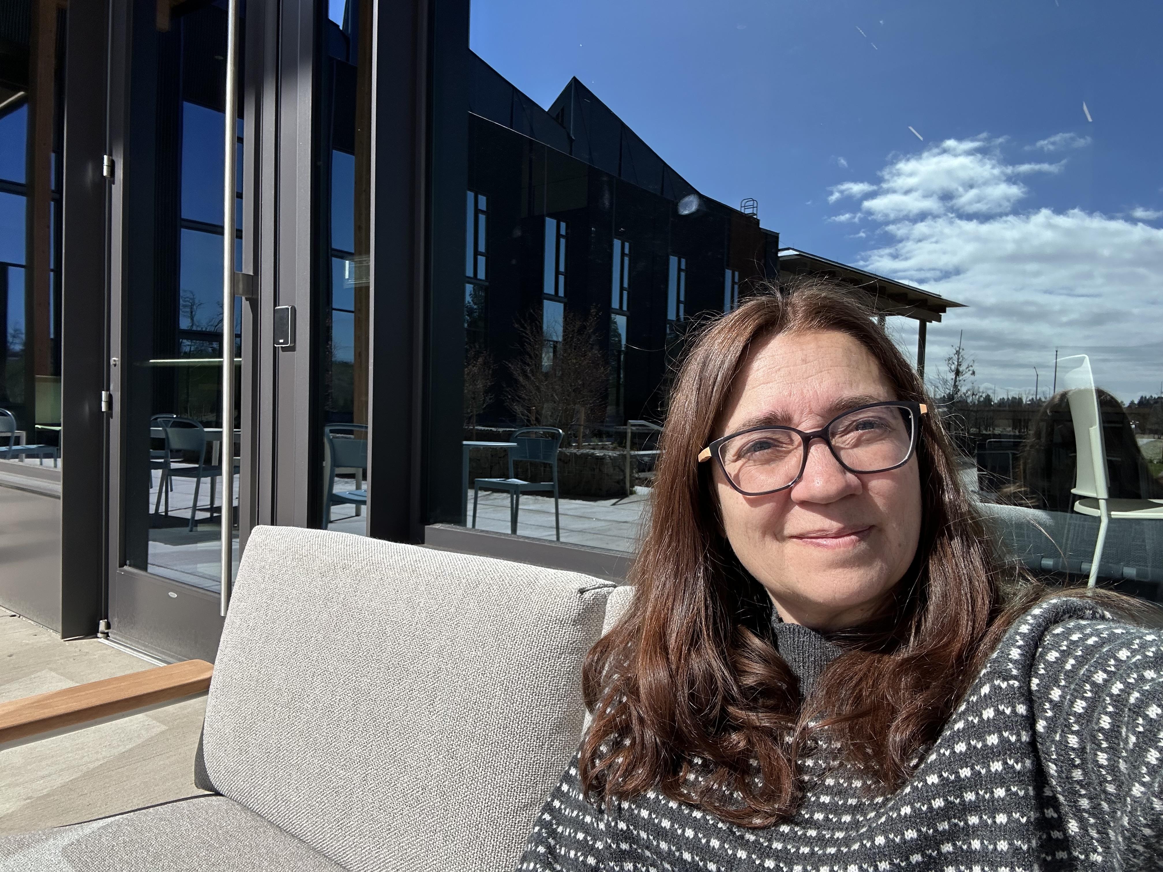 A woman with glasses smiles while sitting outdoors on a sunny day.