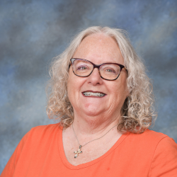 A smiling woman with long white hair, wearing glasses and an orange shirt.