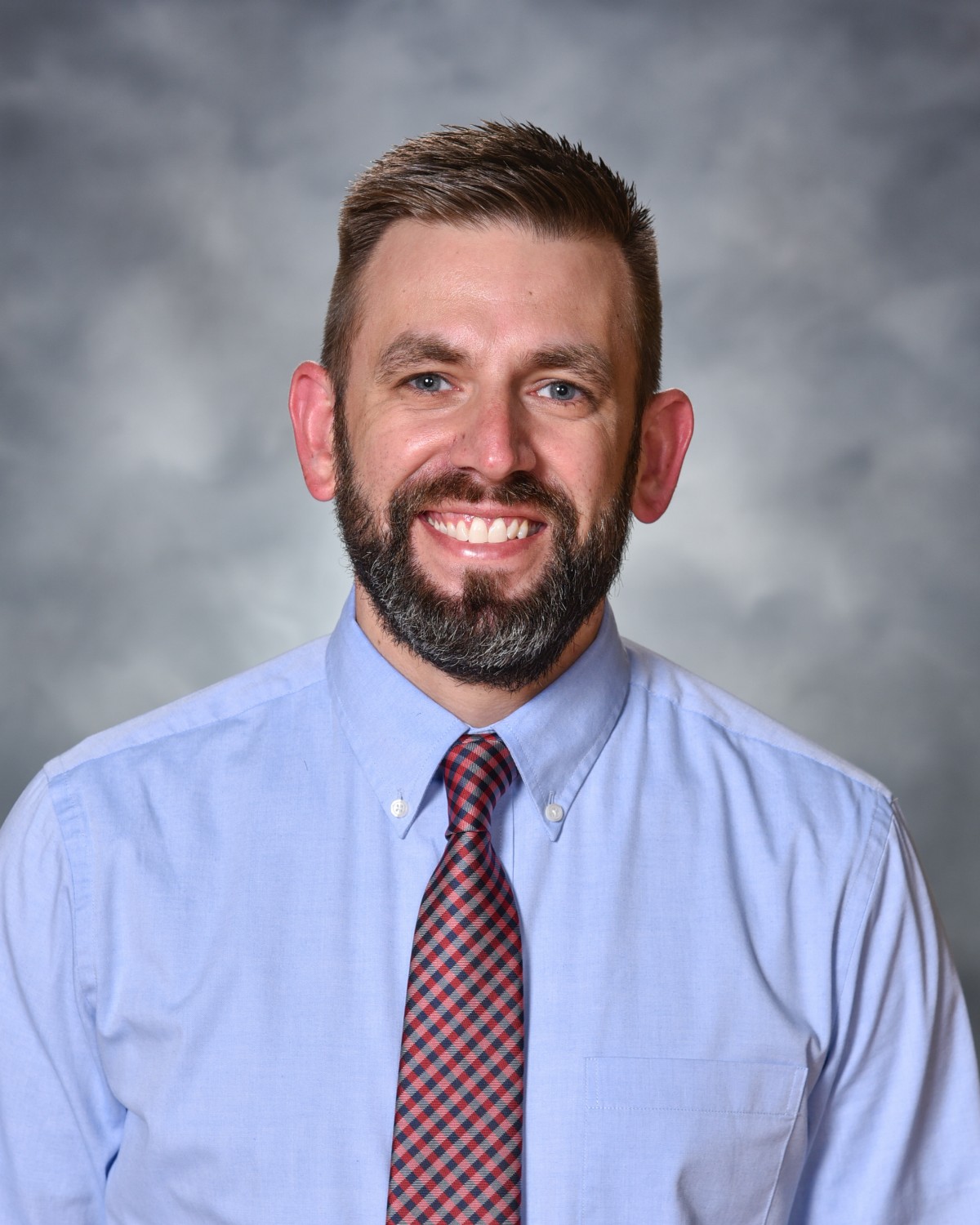 A smiling man with a beard and short hair wears a light blue collared shirt and a striped tie.