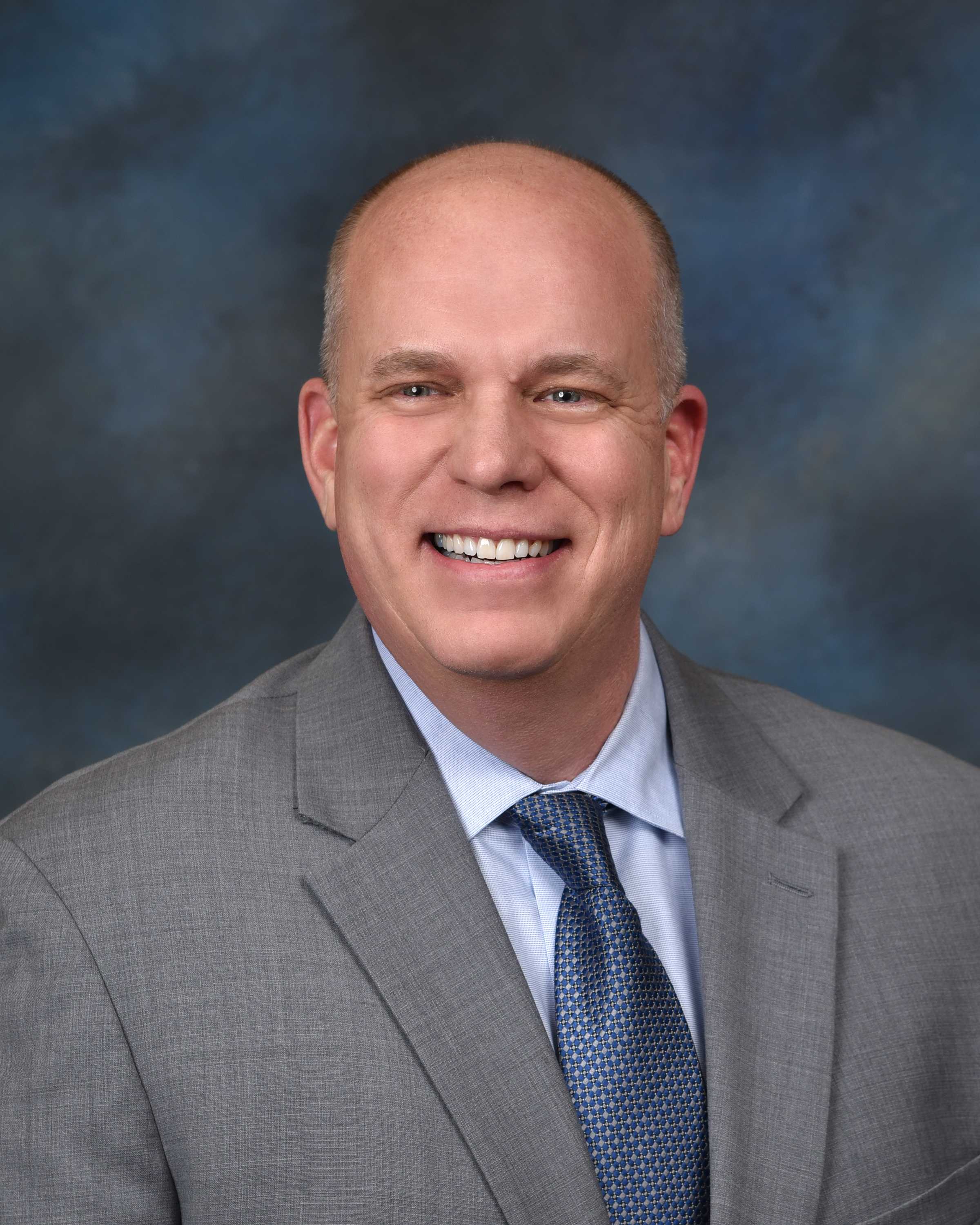 A smiling, man in a grey suit and blue tie poses for a headshot.