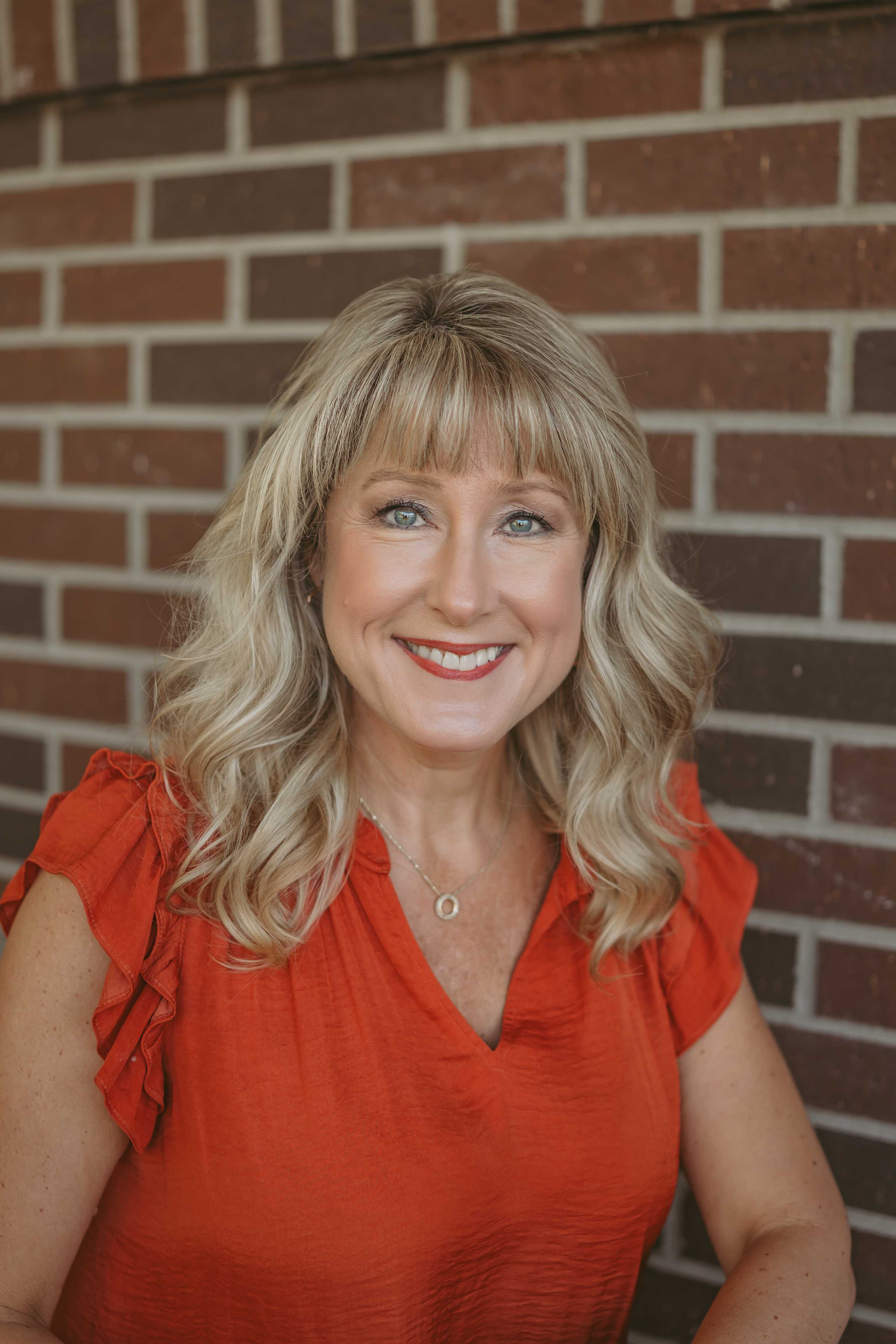 A woman with blonde hair smiles in front of a brick wall.